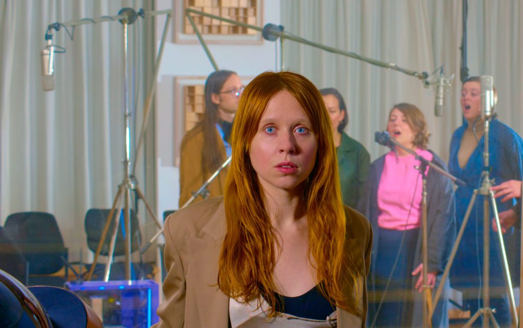 Holly Herndon faces the camera while standing in front of a group of four vocalists who are singing into an assortment of microphones.