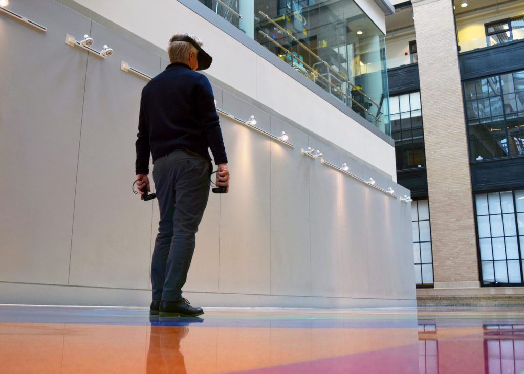 Matthew Ritchie stands on the colorful Sol Lewitt floor at MIT wearing a VR headset and holding hand controls.