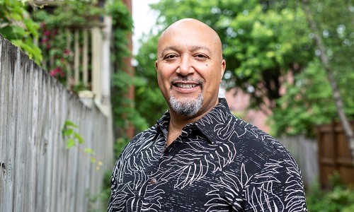 Jason Edward Lewis smiles at the camera while wearing a tropical print collared shirt with lush green trees in the background.